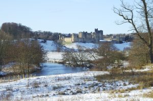 View to Alnwick castle in snow scene with river Aln, blue sky and sunshine.