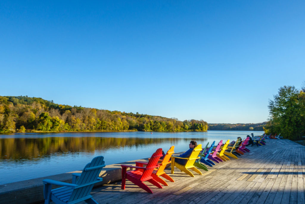 Brightly colored adirondack chairs along a river with trees in background